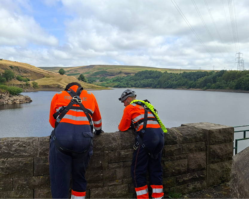 Two men wearing high-visibility overalls stood next to a wall overlooking a large lake